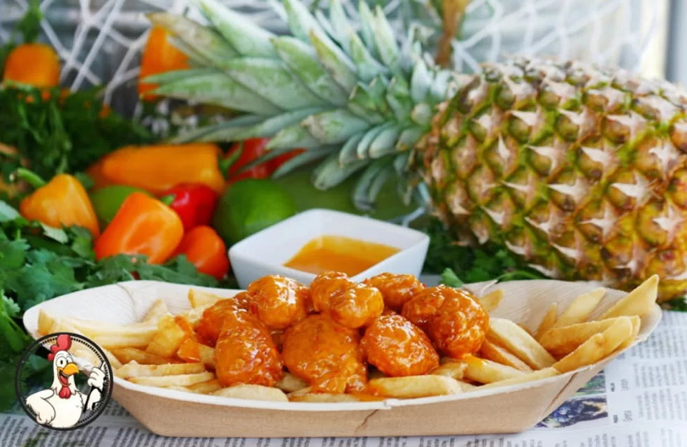 Boom Boom Chicken over fries in a paper boat, a cup of sauce and a pineapple behind